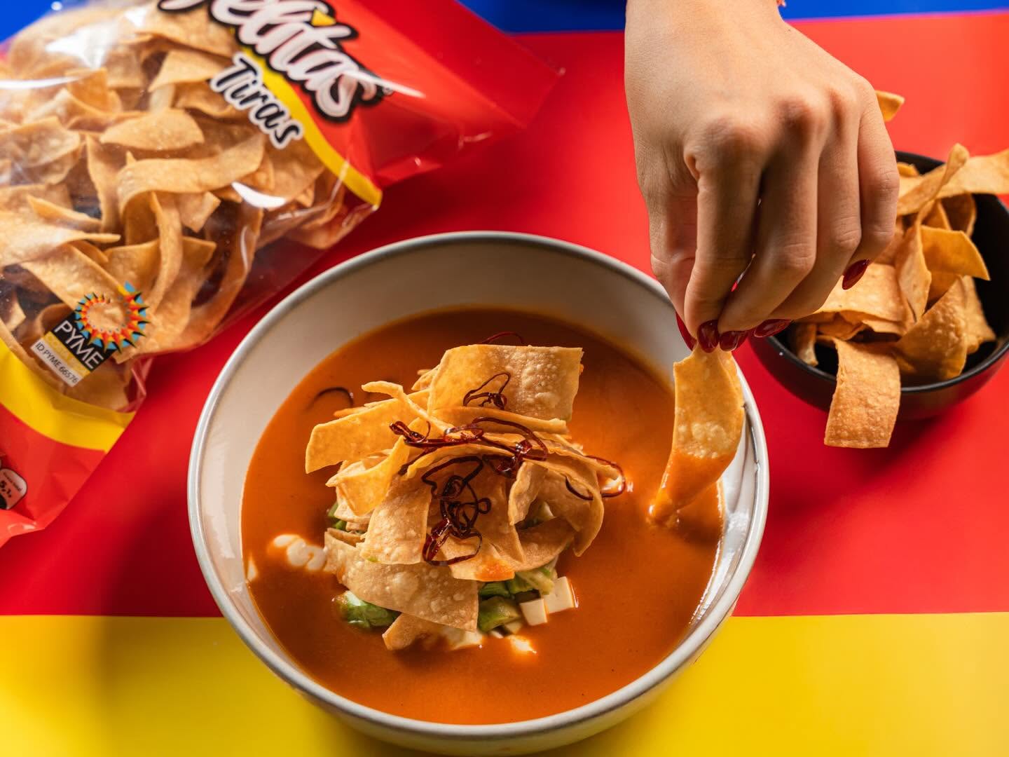 Mano tomando una Rafelita Tiras sobre un tazón de sopa azteca